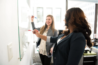 An image of a two women writing on a whiteboard while smiling in the office 