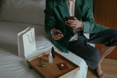 An image of a man using a smartphone while placing order with credit card sitting near cosmetic products on the bed